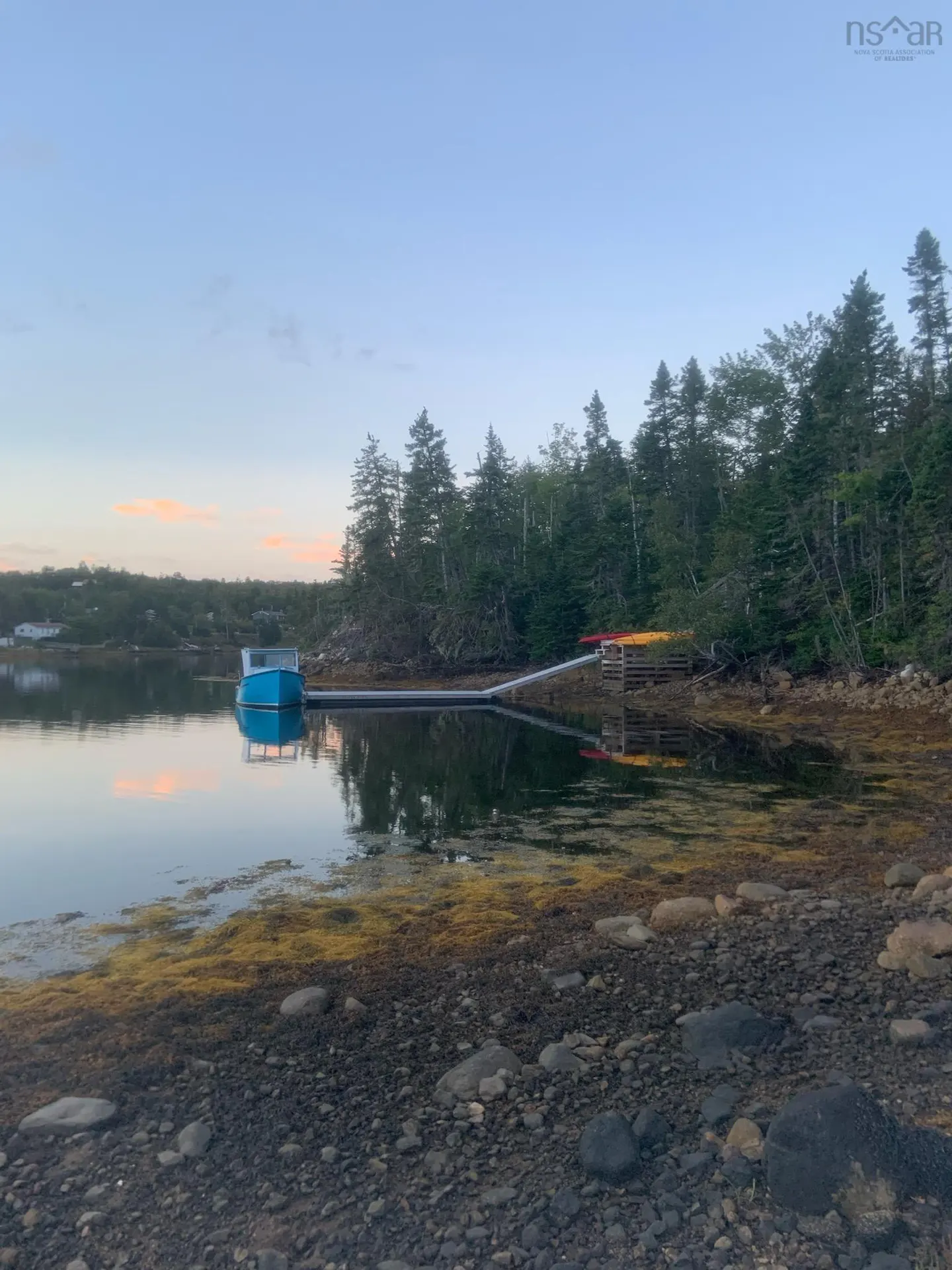 Waterfront view of a blue boat docked at a pier, surrounded by trees and a rocky shoreline at dusk.