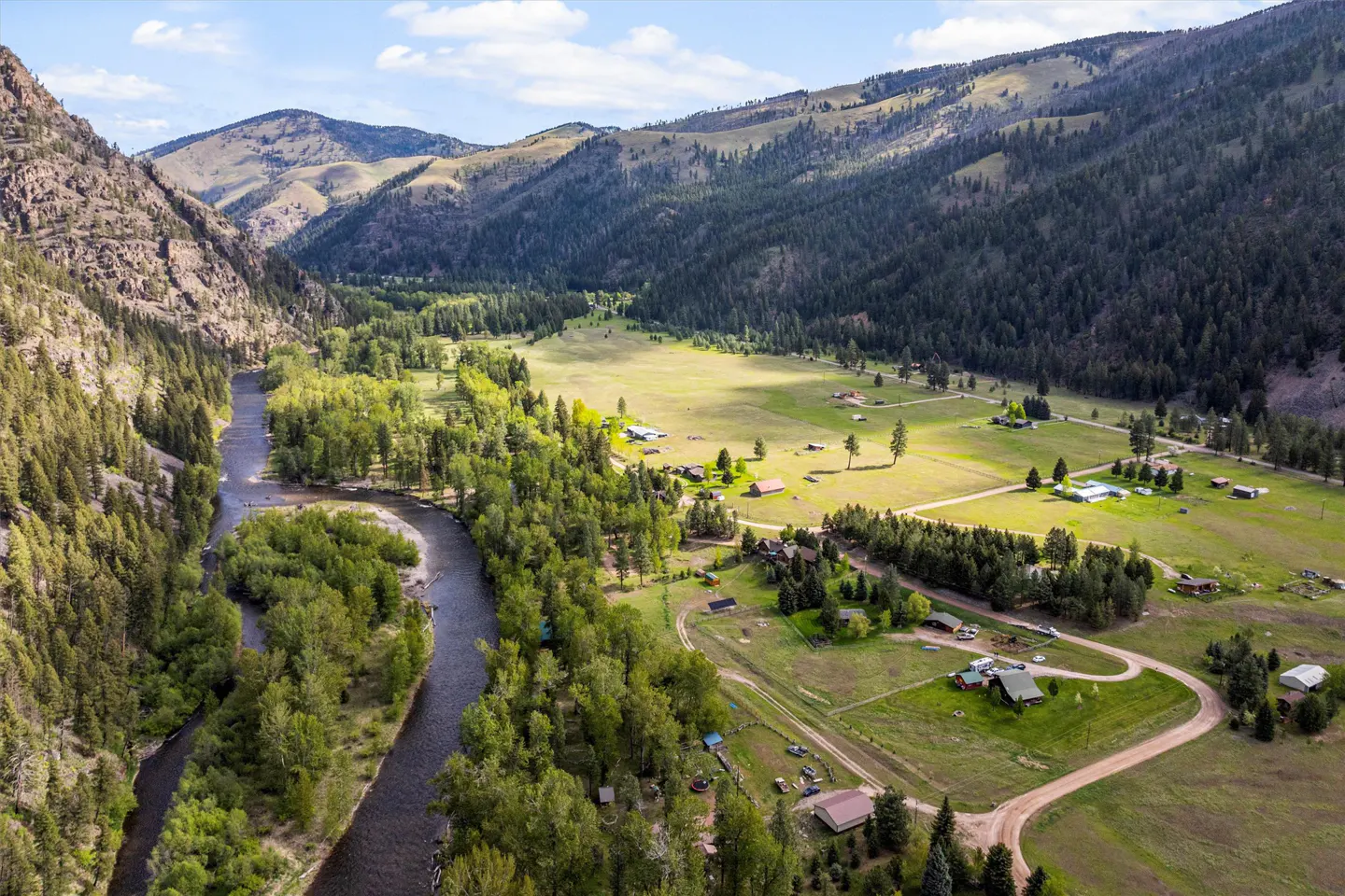 Aerial view of a river valley with green trees, mountains, and scattered houses under a blue sky.