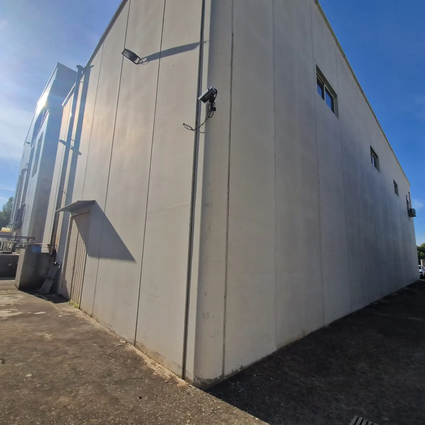 Exterior view of a large, light gray industrial building with a flat roof, small windows, security cameras, and outdoor lighting.