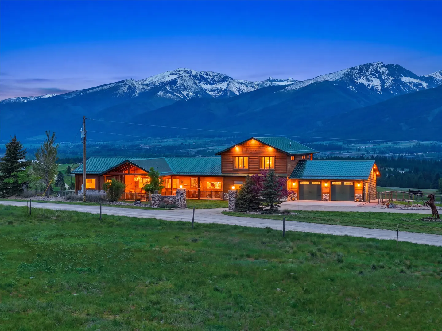 A log home with a green roof sits on a green lawn with snow-capped mountains in the background at dusk.