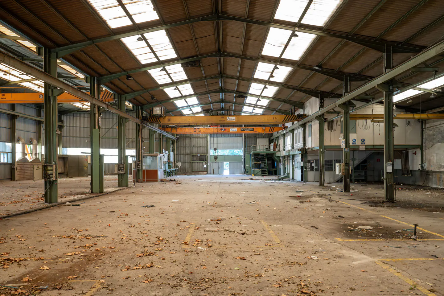 An empty warehouse with a high ceiling, skylights, and a large open door. Yellow cranes hang from the ceiling. The floor is concrete with debris.