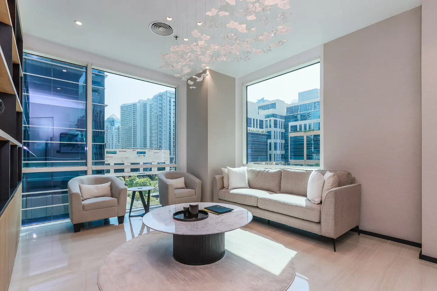 Bright living room with beige sofa, chairs, and round table on a rug. Large windows offer a city view. A modern chandelier hangs above.
