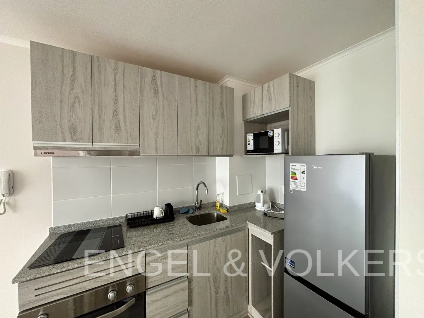 A compact kitchen with wood-grain cabinets, stainless steel appliances, and gray granite countertops. A microwave sits above the counter next to a silver refrigerator.