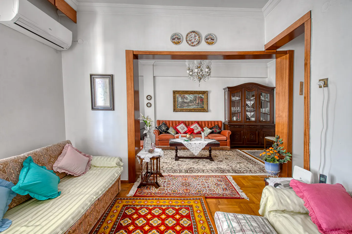 A living room with sofas, rugs, and a wooden cabinet. The walls are white, and there is a wooden frame between the rooms.