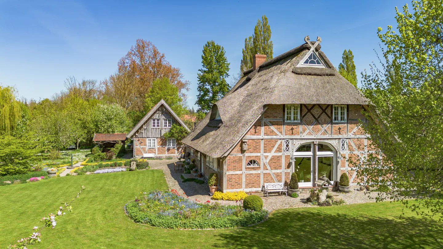 A charming half-timbered brick house with a thatched roof, surrounded by a lush green garden on a sunny day.