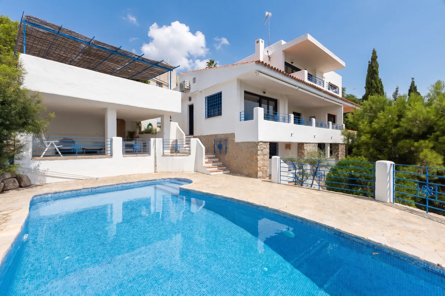 Exterior view of a white two-story house with a blue tiled pool in the foreground on a sunny day.