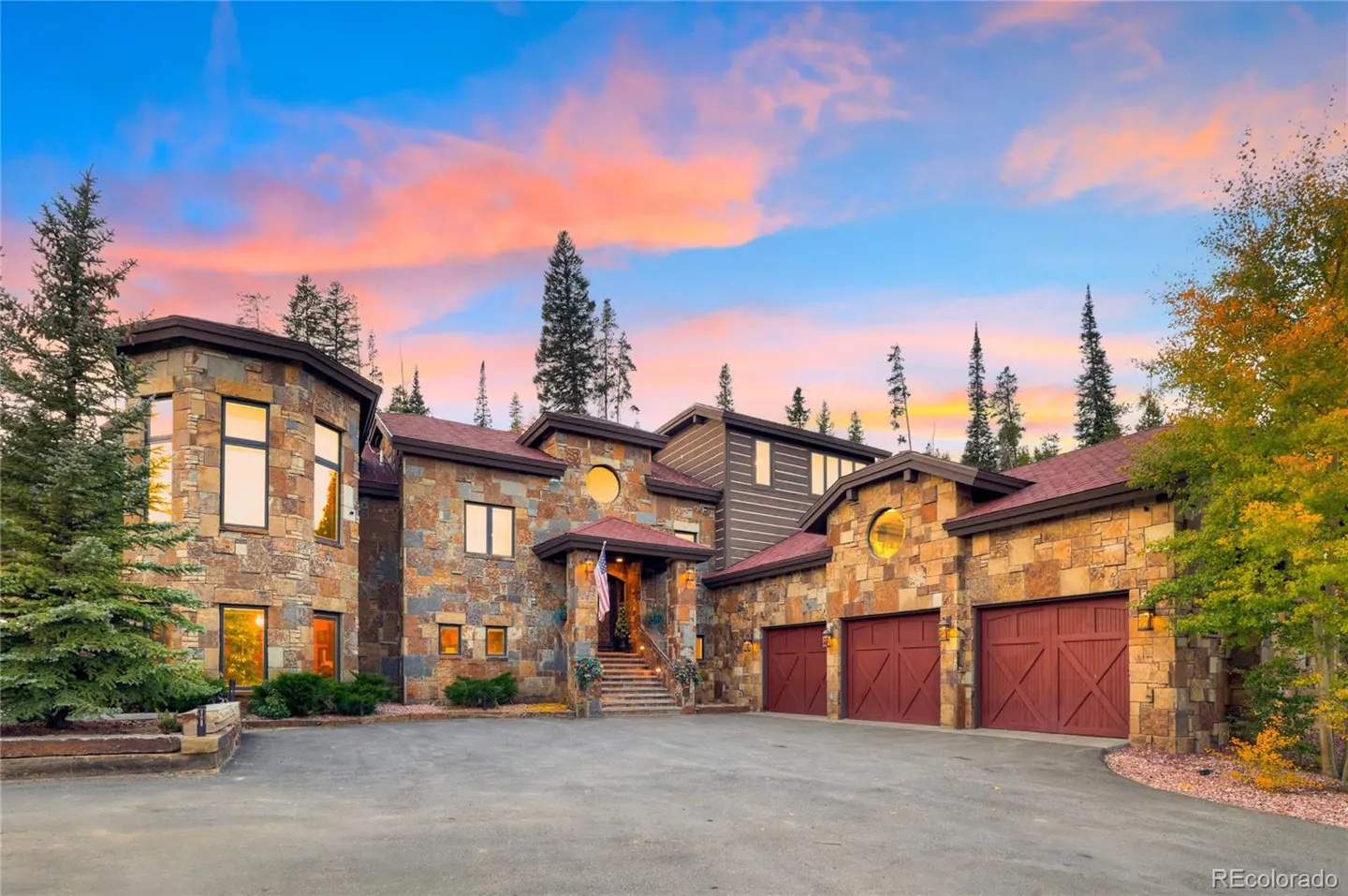 Stone house with red garage doors and roof, set against a colorful sunset sky. Trees surround the property.