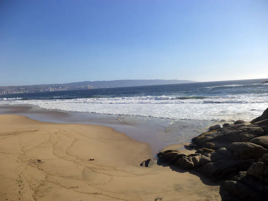 Sandy beach with blue ocean waves and rocks on the right. City skyline visible in the distance under a clear blue sky.