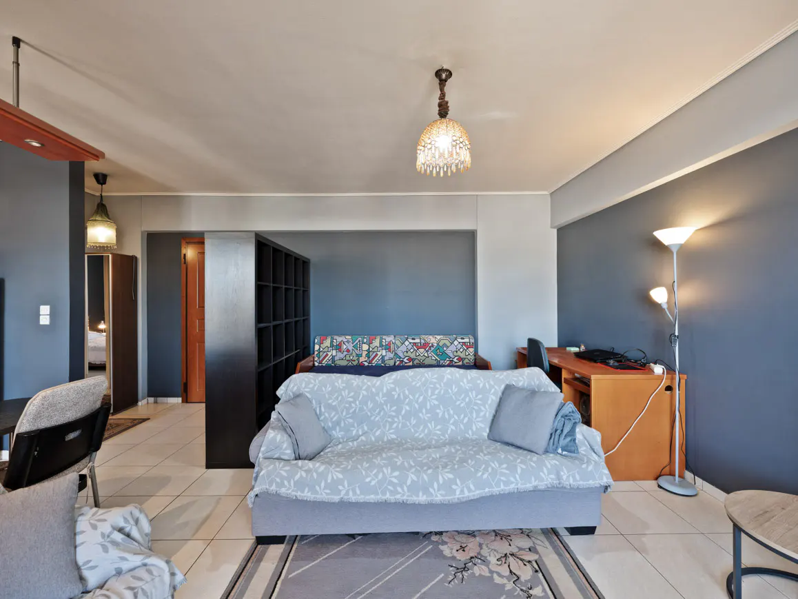 Living room with gray walls, tile floor, and a sofa covered with a light blue floral blanket. A desk, bookshelf, and floor lamp are also visible.