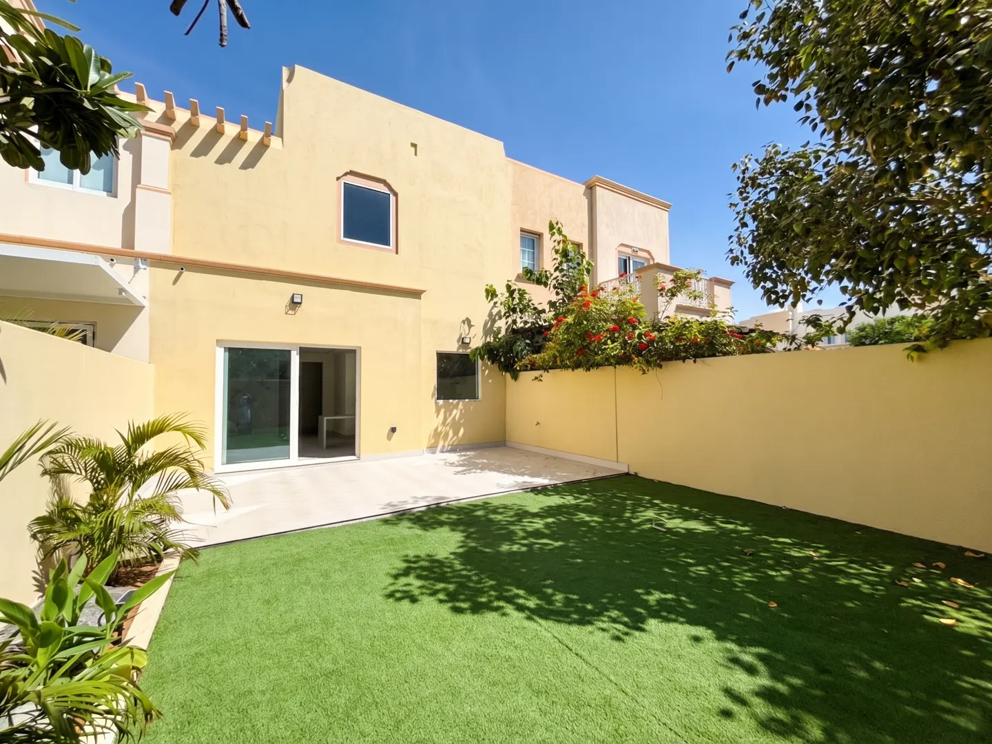 A sunny backyard with green grass, a beige house with a sliding glass door, and a beige wall with red flowers.