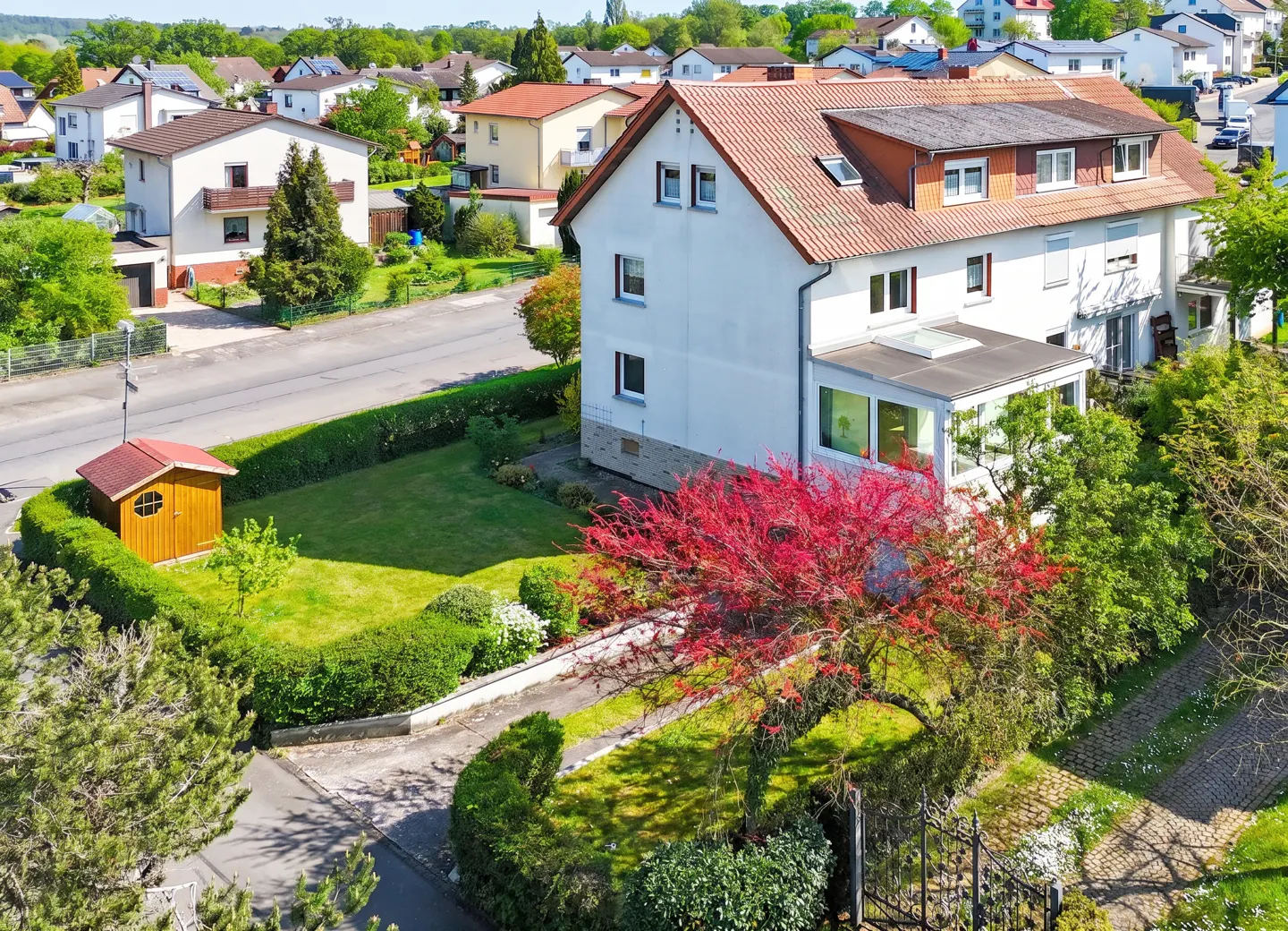 Aerial view of a two-story white house with a red tile roof, surrounded by green lawns, trees, and a small wooden shed.