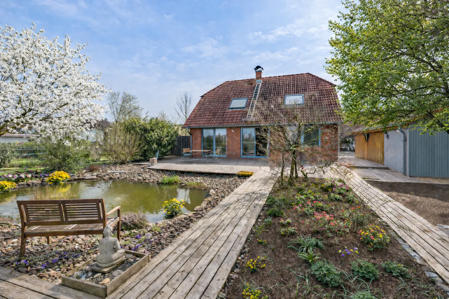 A brick house with a red tile roof overlooks a pond with a wooden bench and walkway in a lush garden.