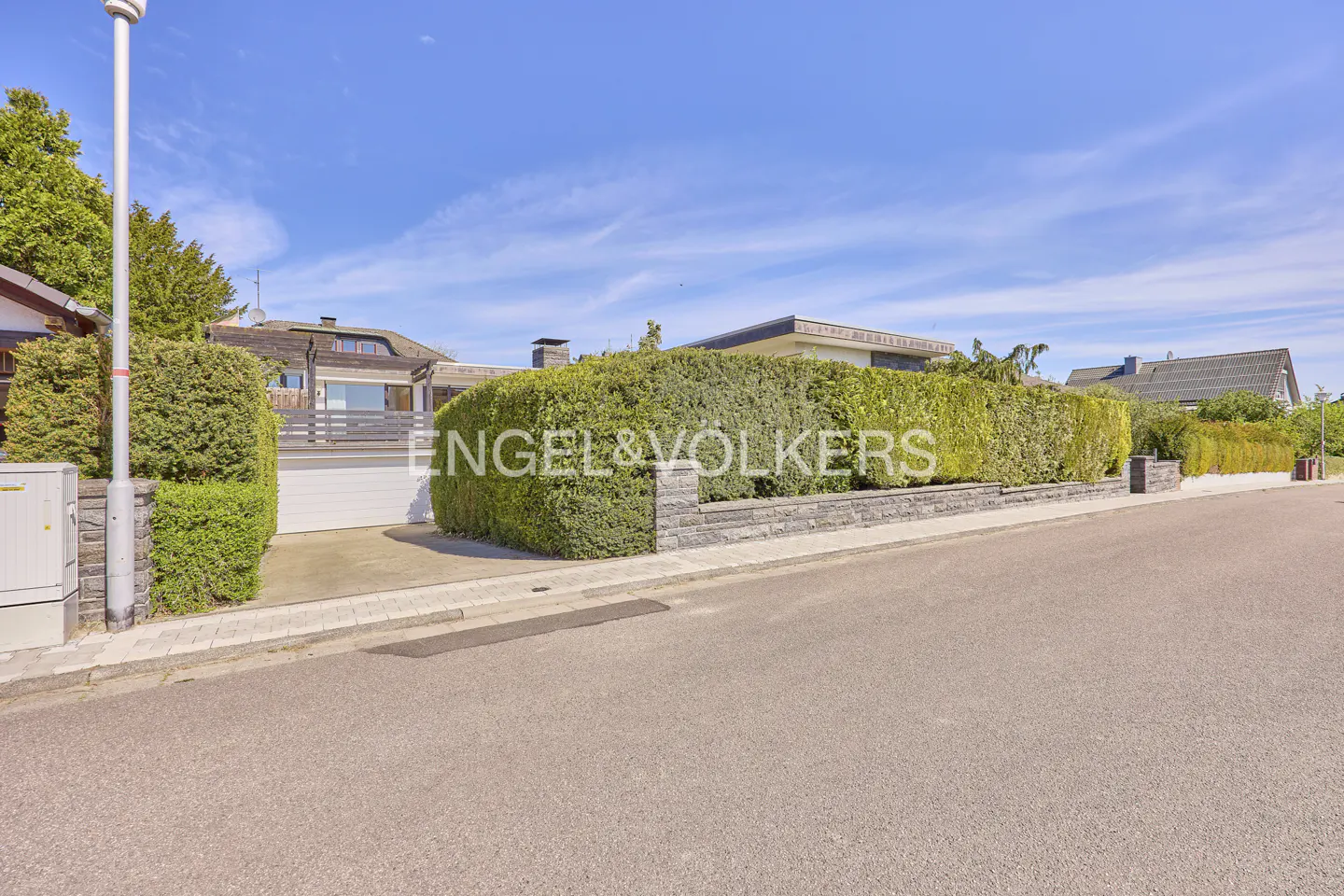 A modern house with a green hedge and a gray stone wall under a blue sky.