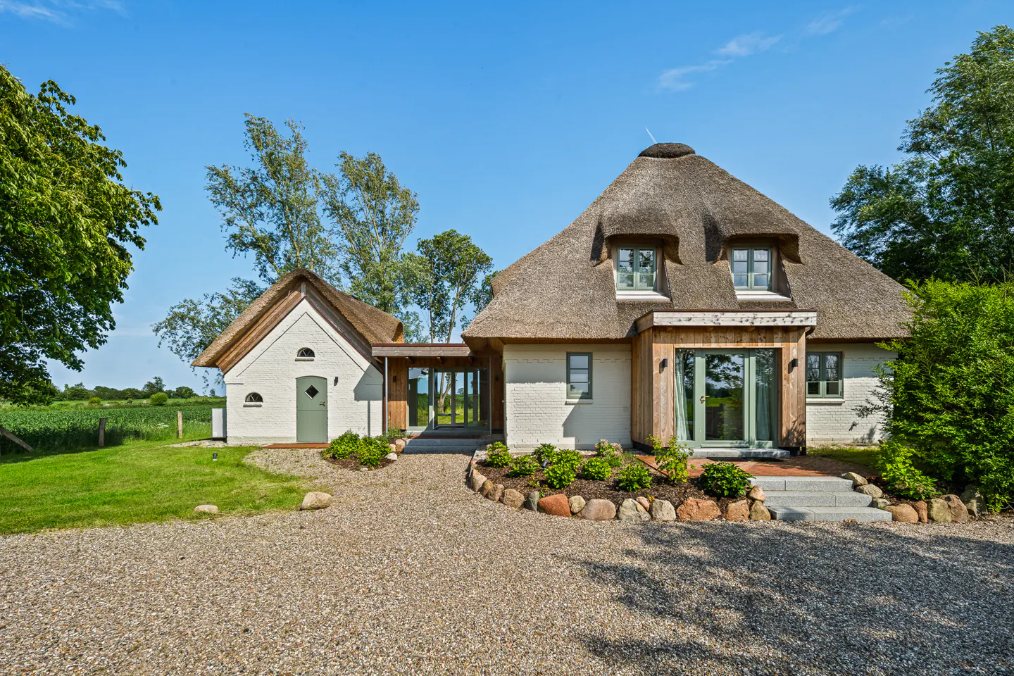 A white cottage with a thatched roof and a gravel driveway, set against a bright blue sky and lush green landscape.