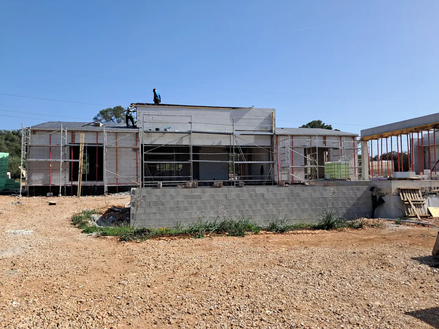 Construction site with a gray block house, scaffolding, and two workers on the roof under a clear blue sky.