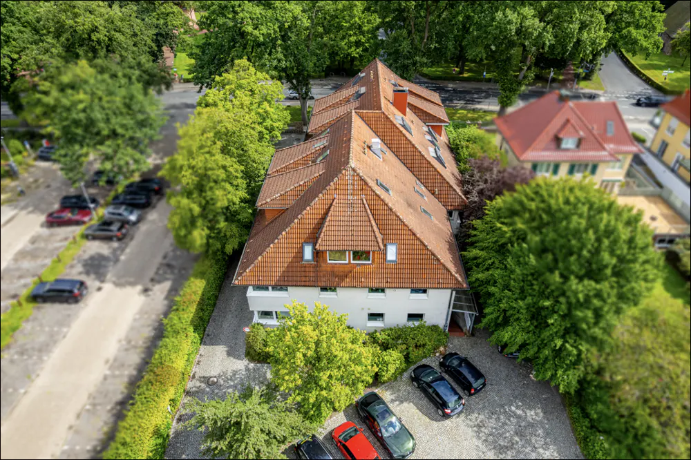 Aerial view of a white building with a brown tiled roof, surrounded by green trees and parked cars.