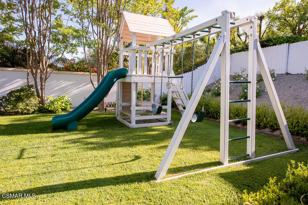 A white wooden playset with a green slide and swings sits on a green lawn in a backyard.