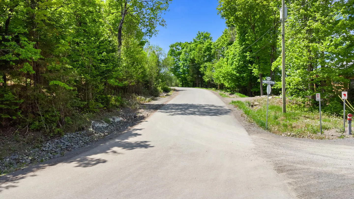 Paved road curves uphill, framed by green trees under a blue sky. Roadside signs and utility poles are visible.