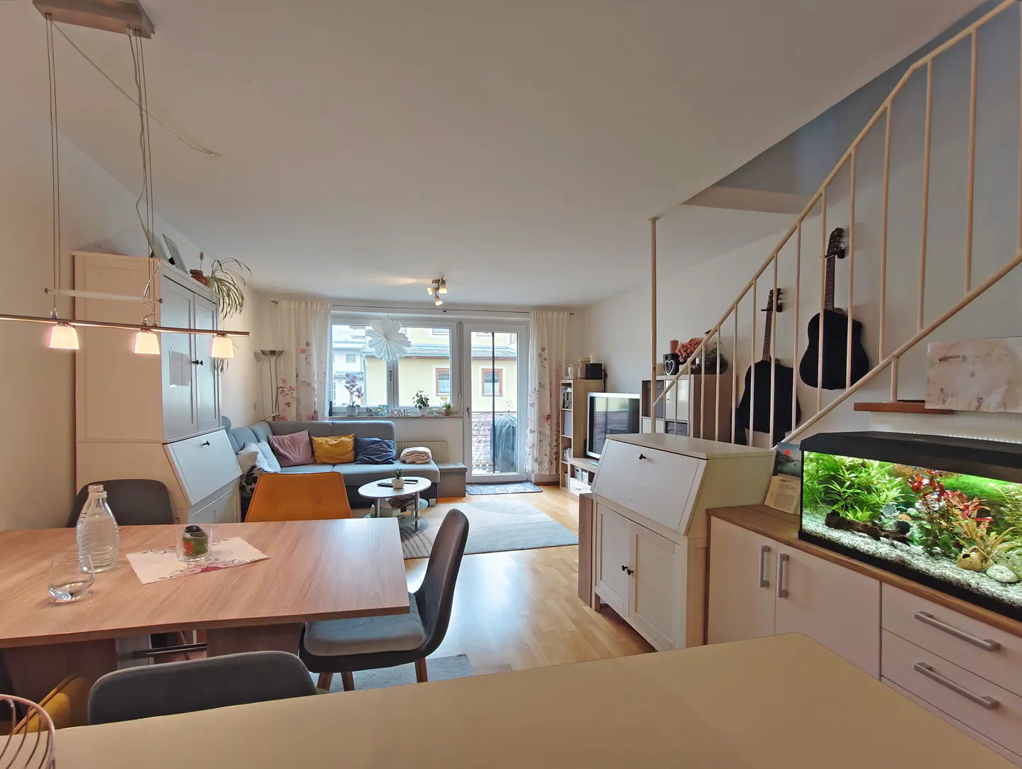 A bright living room with a dining table, gray sofa, and an aquarium under the stairs. Two guitars hang on the wall.