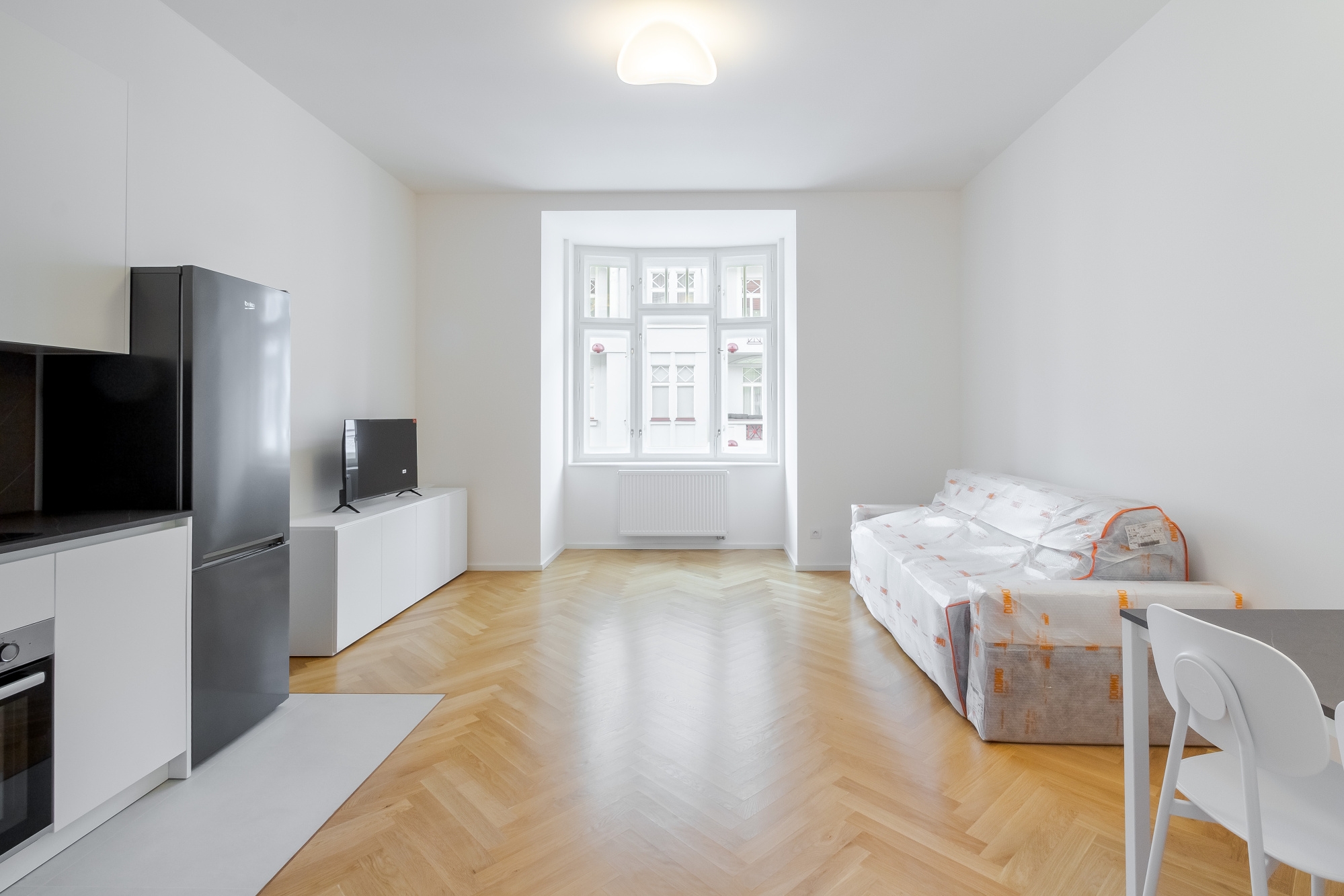 Bright studio apartment with herringbone wood floors, a black refrigerator, a sofa covered in plastic, and a bay window.