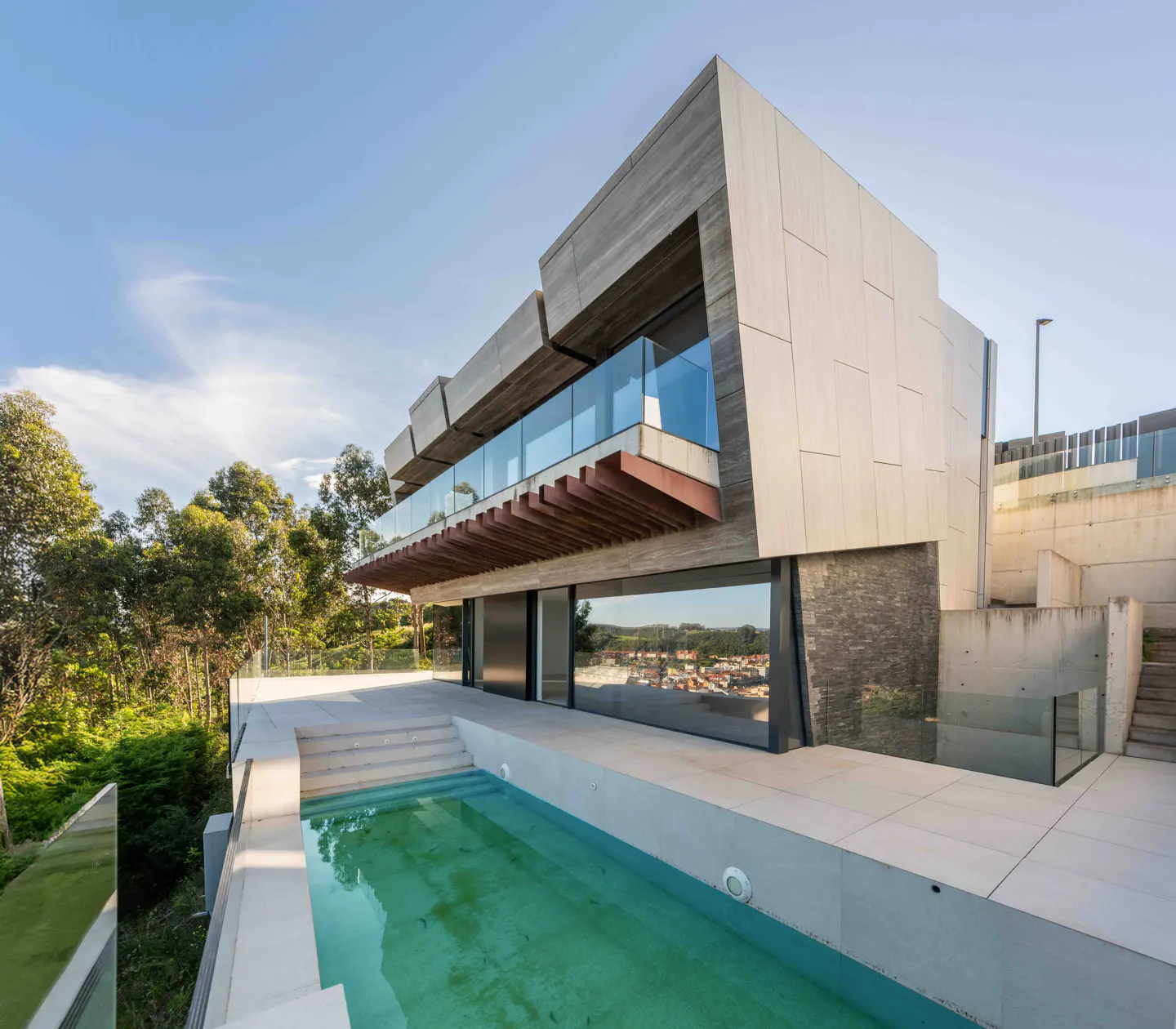 Modern multi-level home with a swimming pool, glass balcony, and stone accents, set against a backdrop of trees and sky.