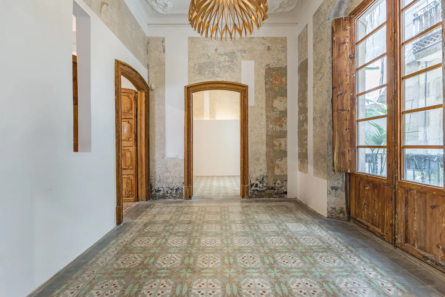 A room with patterned tile floors, wooden doors, and large windows. Walls are a mix of white and distressed plaster. A modern wooden chandelier hangs from the ceiling.
