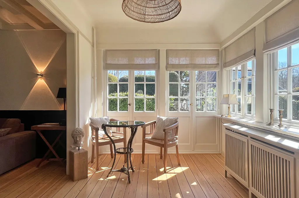 Sunlit room with wood floors, white walls, and a table with two chairs. Windows with beige blinds overlook a green garden. A bust sits on a wooden stand.