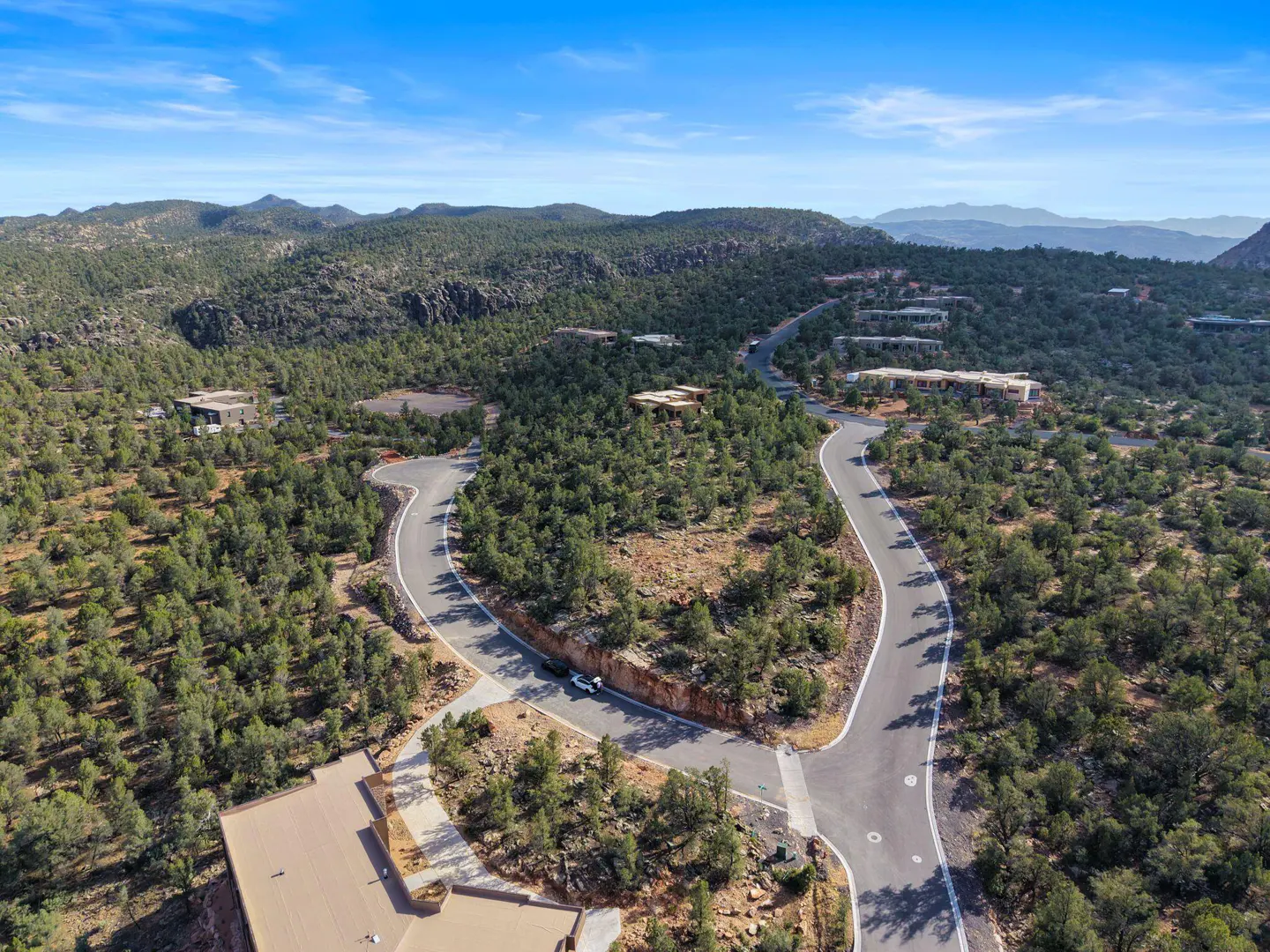 Aerial view of a winding road through a green, mountainous landscape with scattered houses under a blue sky.