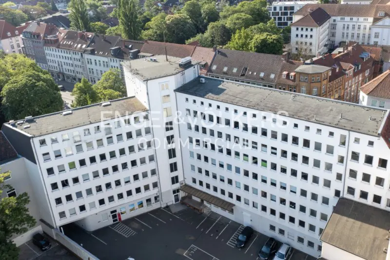 Aerial view of a white, multi-story commercial building with a parking lot and surrounding trees and buildings.