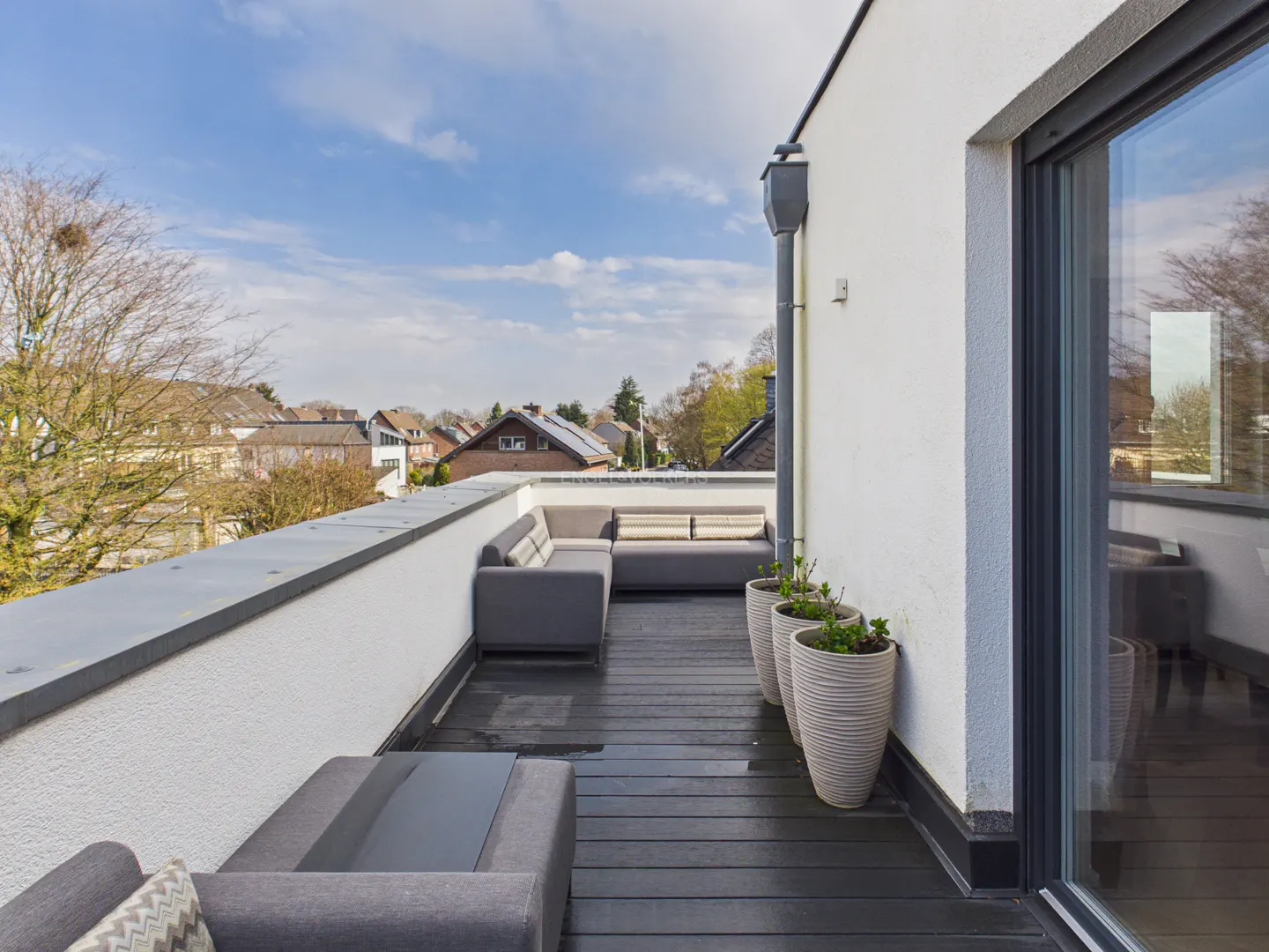 A modern rooftop terrace with gray outdoor furniture, potted plants, and a dark wood floor, overlooking a residential neighborhood under a blue sky.
