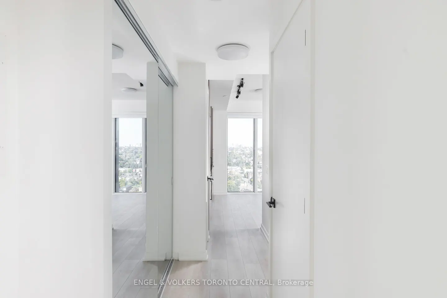 Bright, white hallway with light wood floors, sliding mirrored closet doors, and a view of the city through windows.