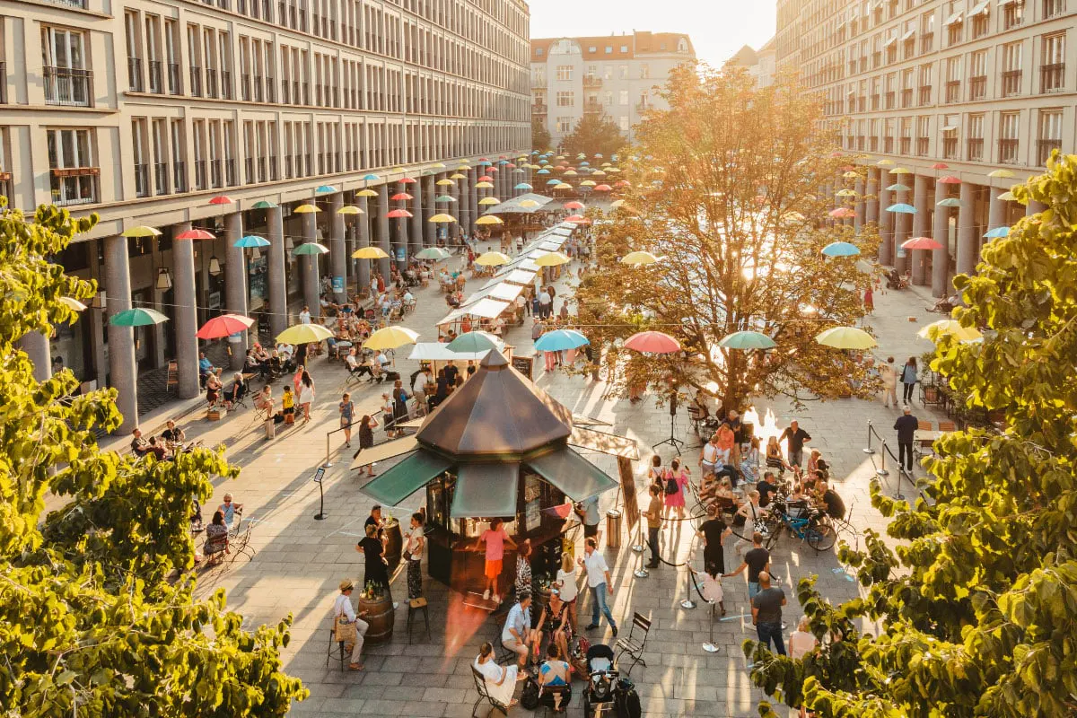 Outdoor market scene with colorful umbrellas strung between buildings, people, and a central kiosk.