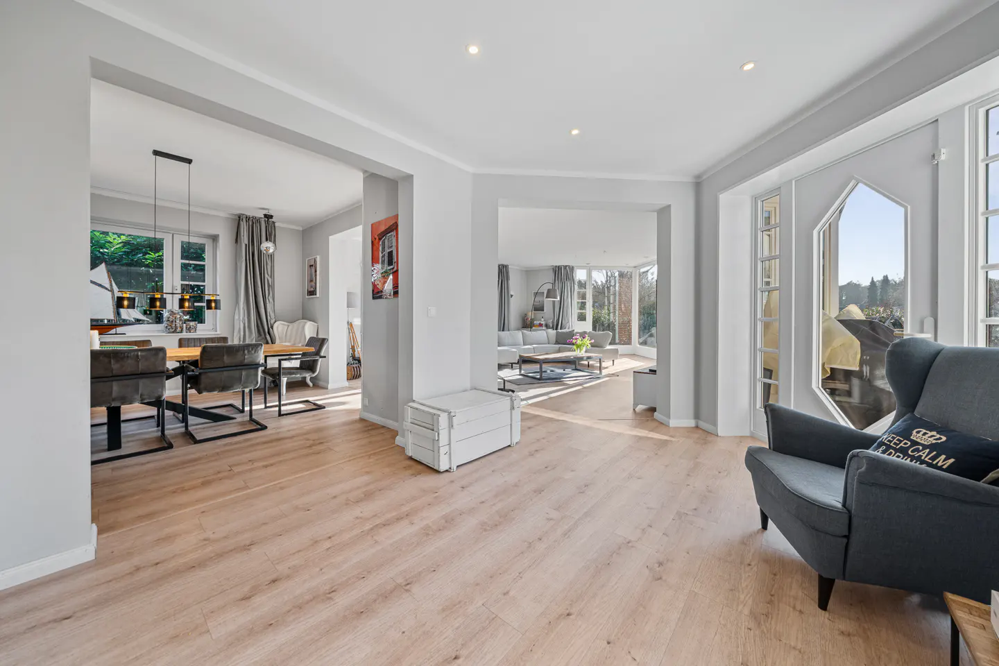 Open-plan living area with light wood floors, a grey armchair, a dining room, and a bright, modern lounge in the background.