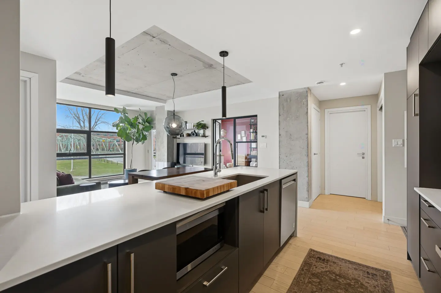 Modern kitchen with white countertops, dark cabinets, and stainless steel appliances. A large window overlooks a green space.