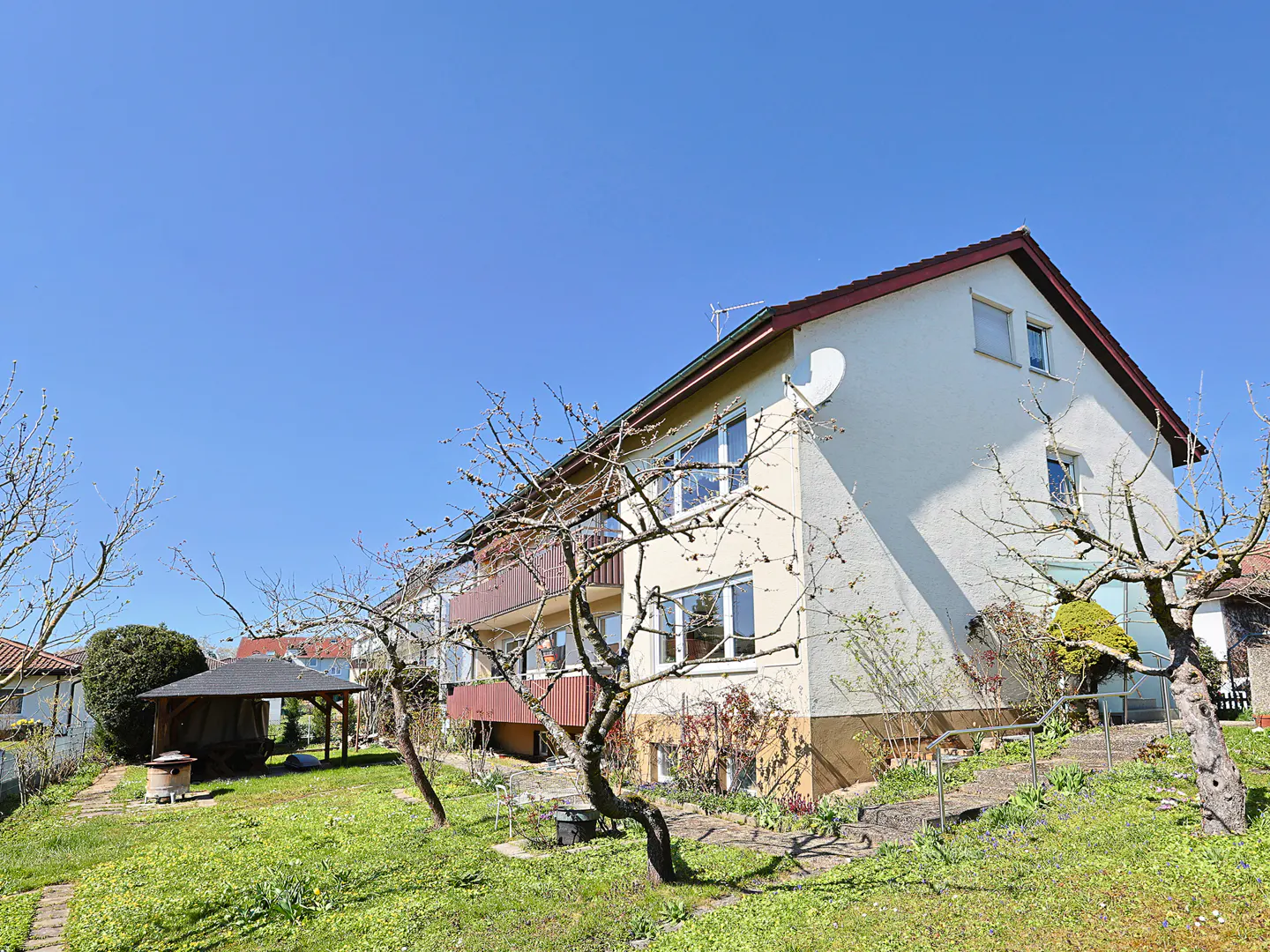 Two-story house with a red roof and a brown balcony, surrounded by a green lawn and trees under a clear blue sky.