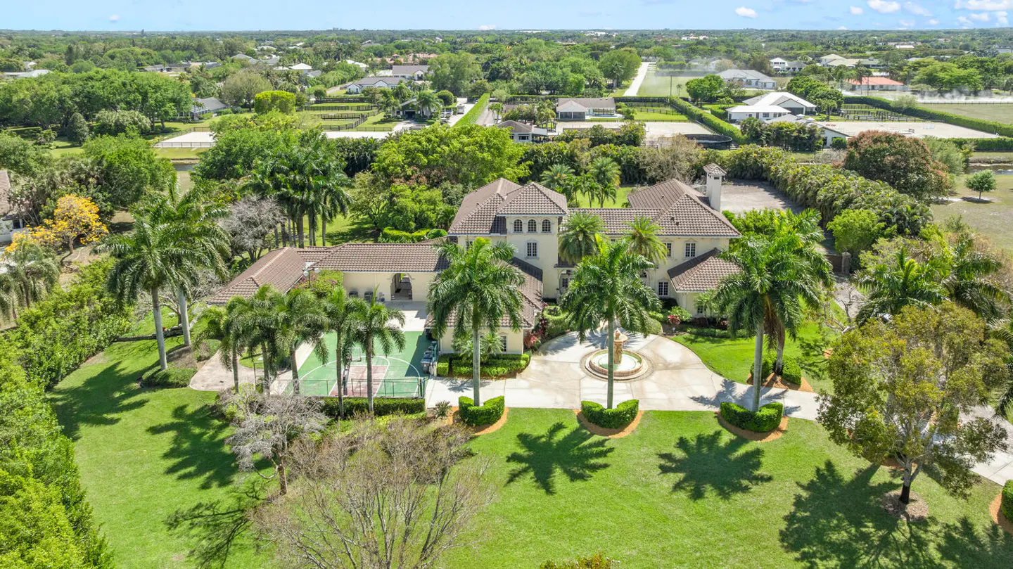 Aerial view of a large, beige mansion with a brown tile roof, palm trees, a circular driveway with a fountain, and a green basketball court.