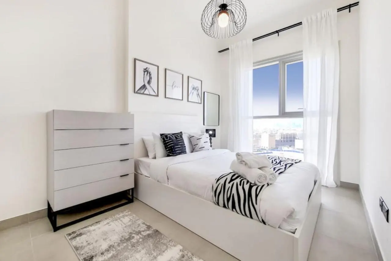 Modern bedroom with white bed, zebra-print accents, grey dresser, and large window with city view under a wire pendant light.