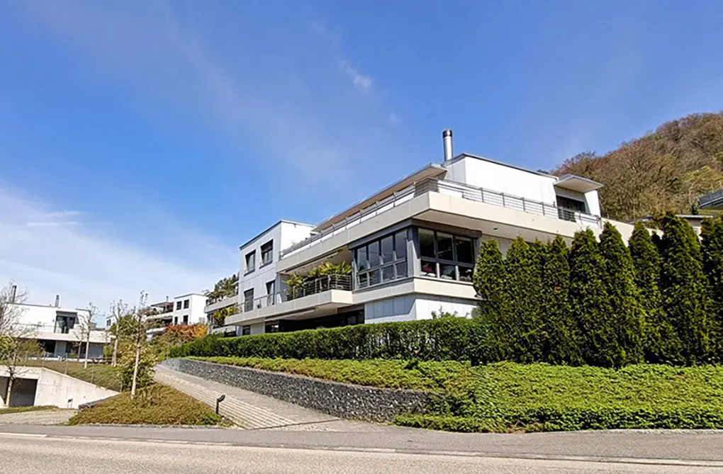 Modern white apartment building with balconies, surrounded by green hedges and trees under a blue sky.