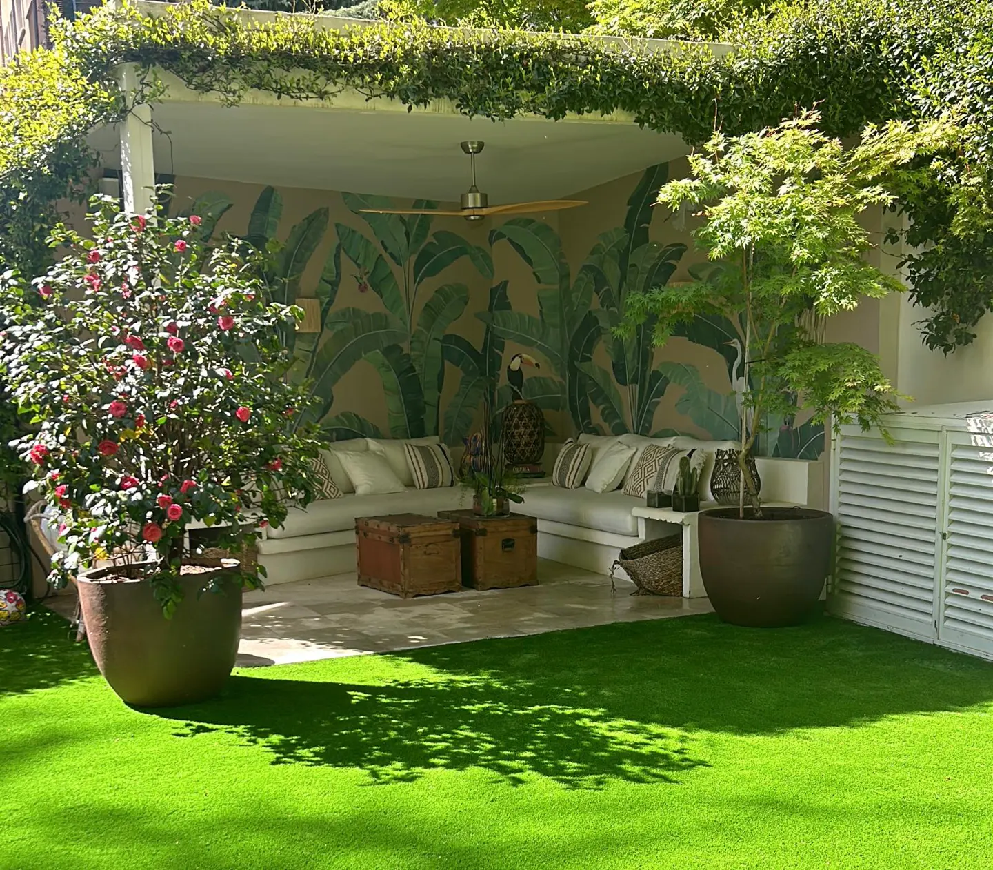 Covered patio with white sectional seating, tropical leaf wallpaper, ceiling fan, and potted plants on a green lawn.
