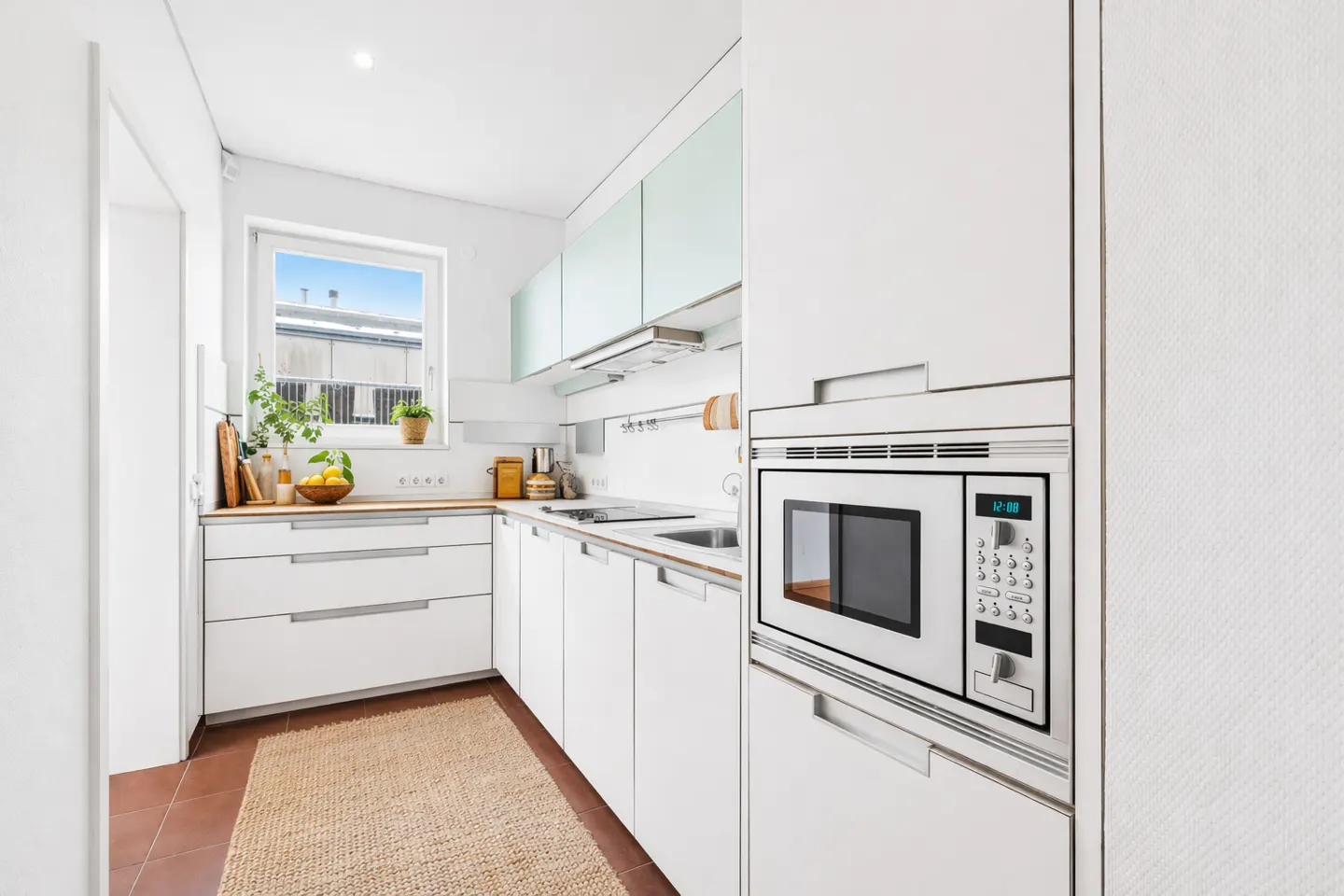 Bright kitchen with white cabinets, wood countertops, and a built-in microwave. A woven rug sits on the brown tile floor.