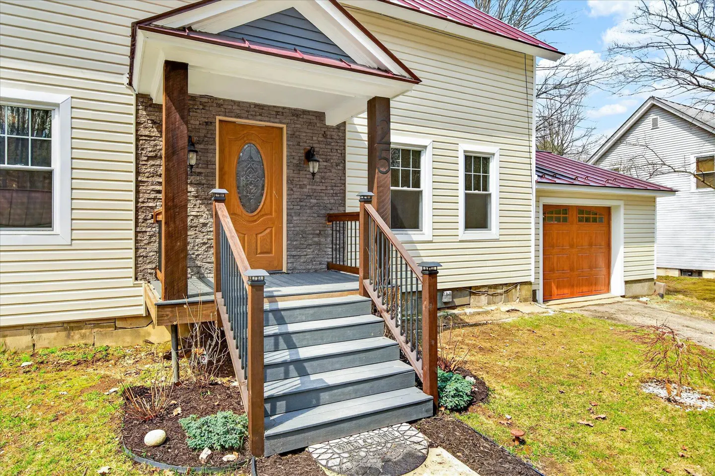 Tan house with a red roof, stone accents, and a wooden door. Gray steps lead to the front door with wooden railings. A garage is visible on the right.