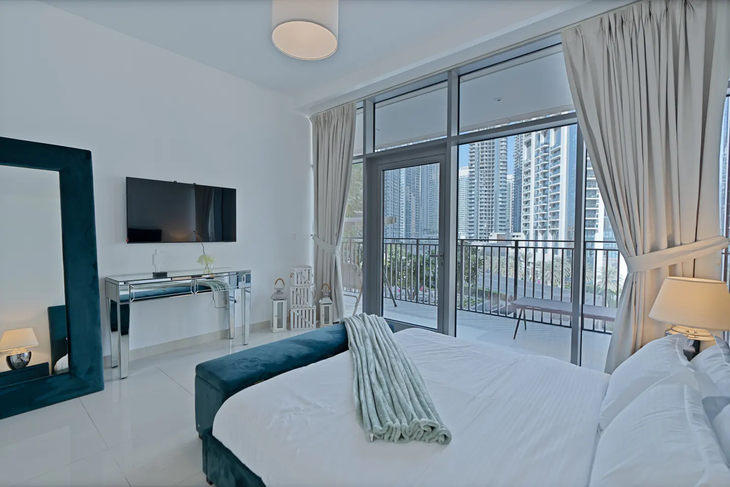Bright bedroom with white linens, blue accents, and a balcony view of city skyscrapers. A large mirror and TV are on the wall.