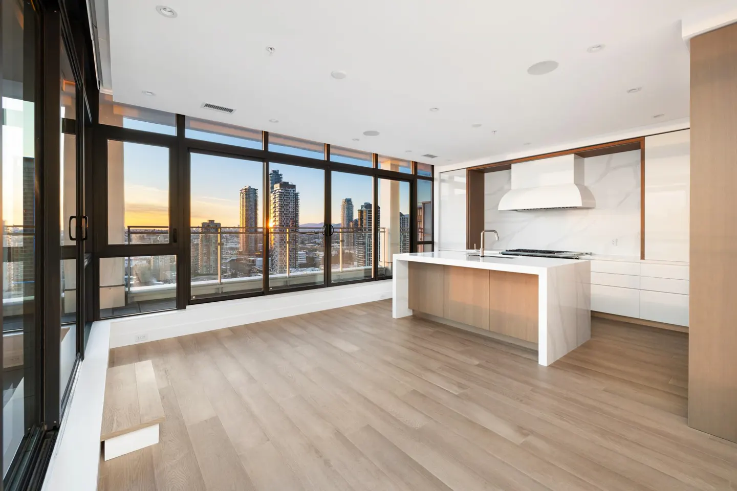 Modern kitchen with white island, light wood floors, and floor-to-ceiling windows overlooking a city skyline at sunset.