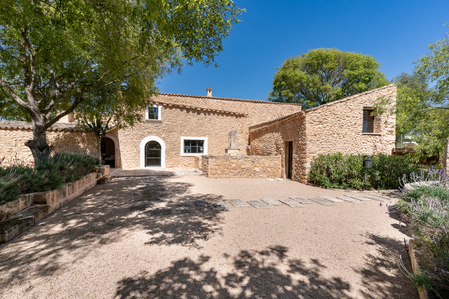Exterior view of a tan stone house with a gravel driveway and green trees under a blue sky.