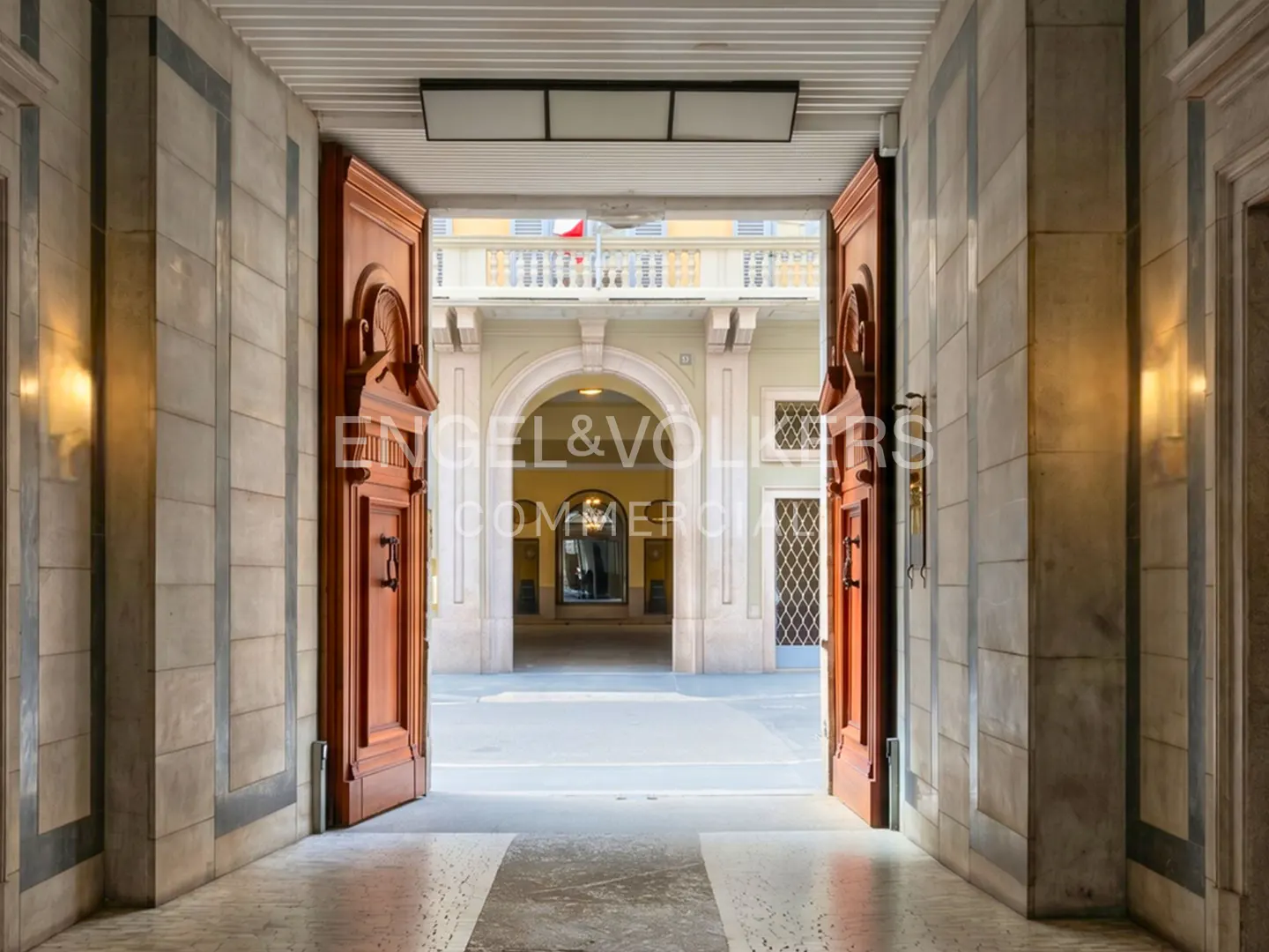 View through open wooden doors into a courtyard with arched walkways and a flag on a building in the background.