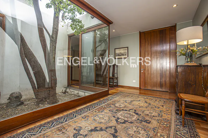 Interior view with a tree in a glass-enclosed courtyard, wood floors, and an ornate rug. A wooden door and furniture are visible.