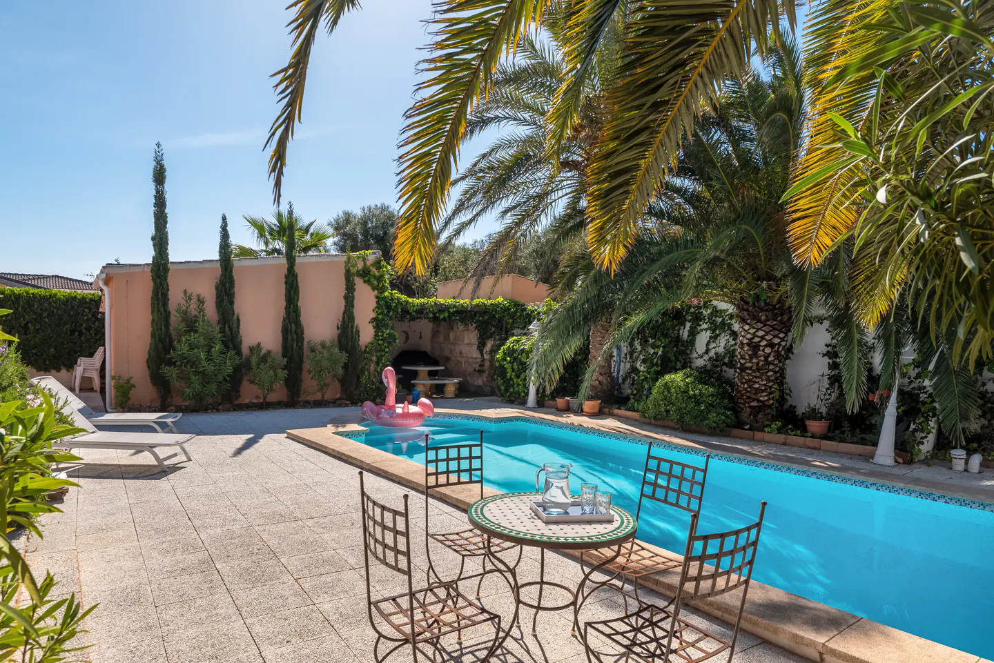 Sunny backyard with a blue swimming pool, palm trees, patio furniture, and a pink flamingo float under a clear blue sky.