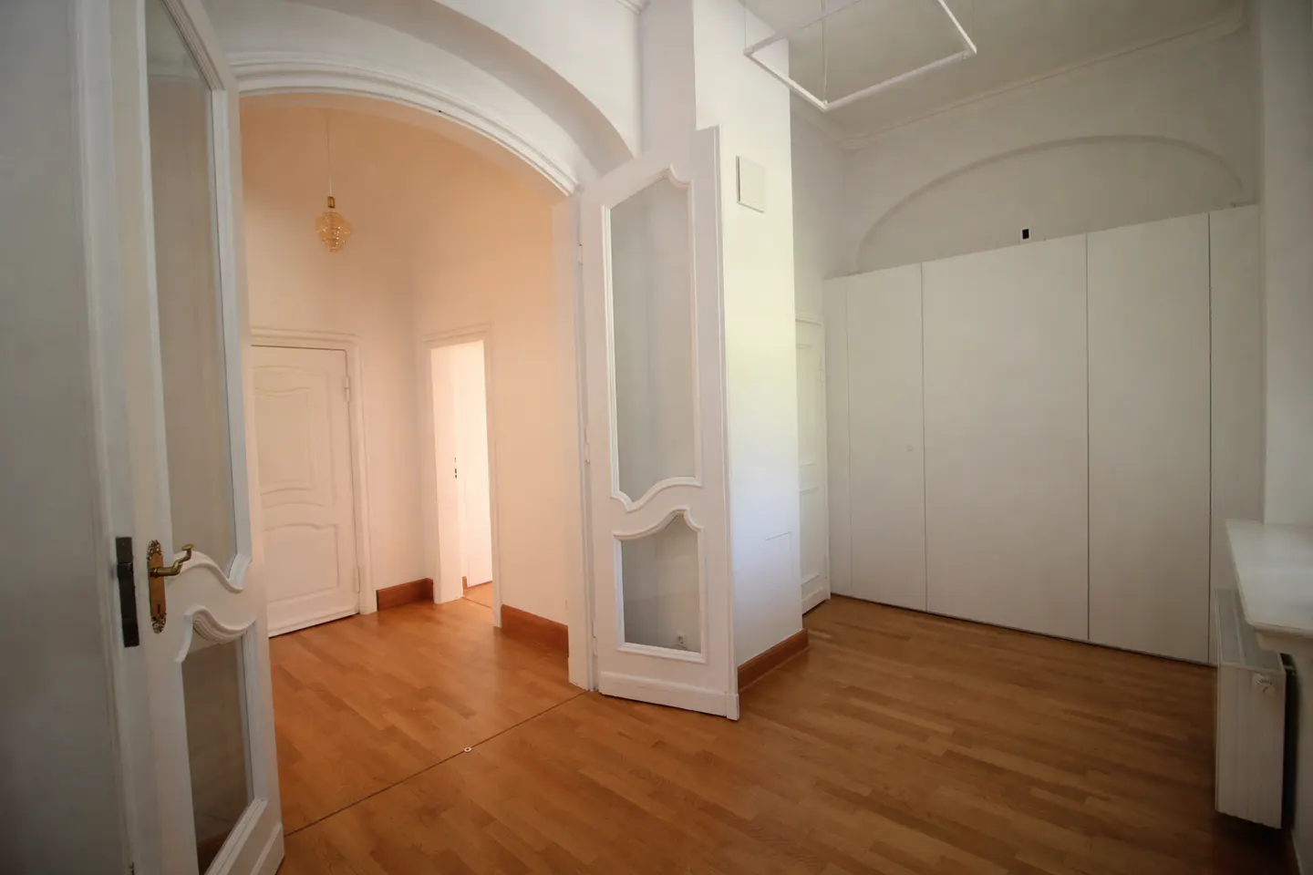 Bright, empty room with hardwood floors, white walls, and arched doorways. A partially open glass door leads to another room.