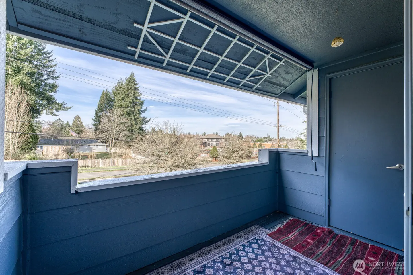 A blue balcony with a view of trees and houses. A patterned rug is on the floor. A blue door is on the right.
