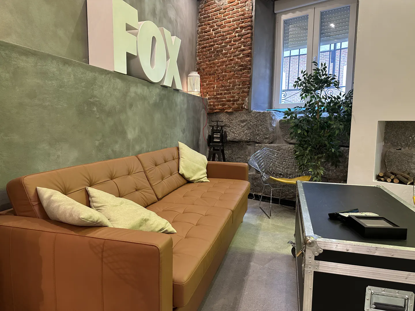 Interior view of a room with a brown leather sofa, light green pillows, and a large "FOX" sign on the wall. A window and a plant are in the background.